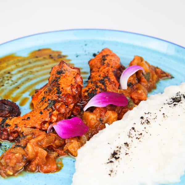 Grilled octopus with a rich red sauce, white rice, and purple flower petals on a blue plate.