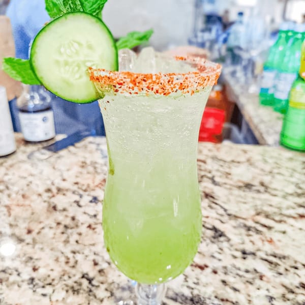 A green Mexican cocktail with a chili-salted rim, garnished with a cucumber slice and mint.