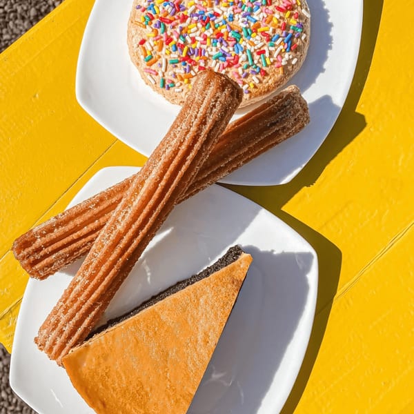 Two plates of Mexican desserts on a bright yellow table, including churros, a sprinkle donut, and a slice of cake.