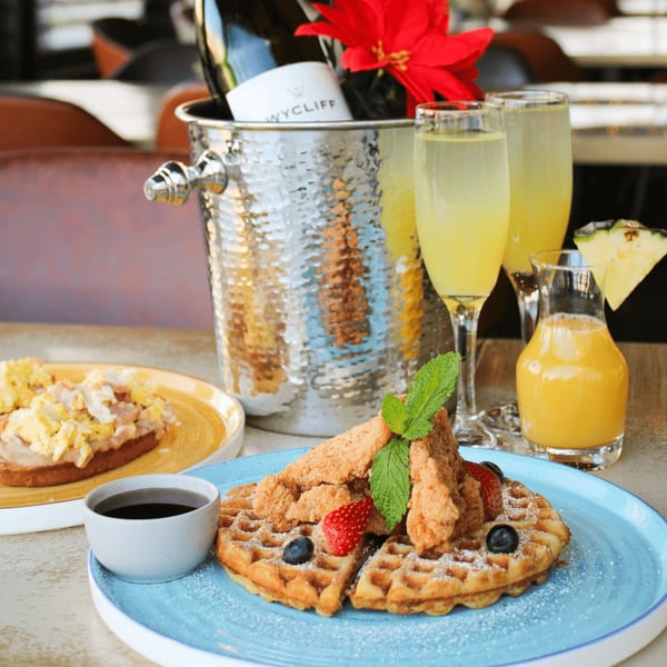 Chicken and waffles, scrambled eggs, mimosas, and champagne in an ice bucket on a table.