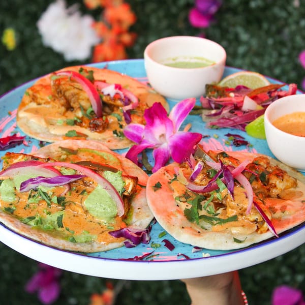 Three vibrant Mexican tacos with red onion, green sauce, and orange sauce on a blue plate.