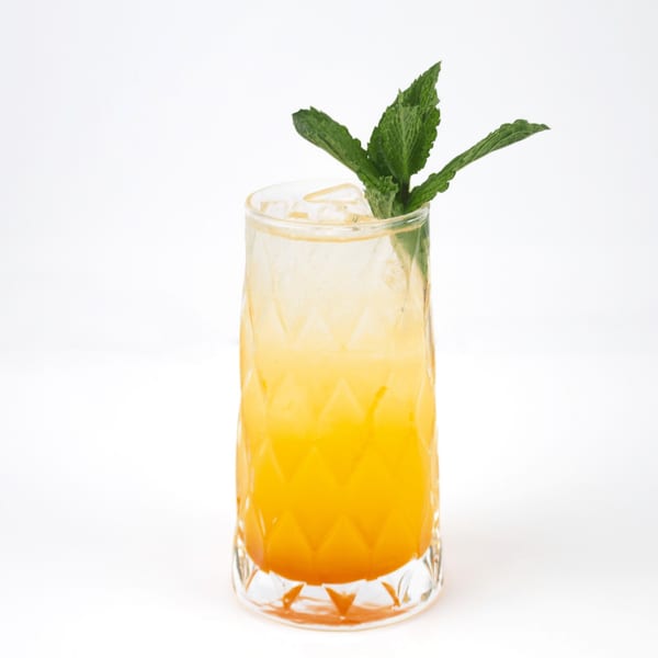 A tall glass of orange drink with ice and a mint garnish on a white background.
