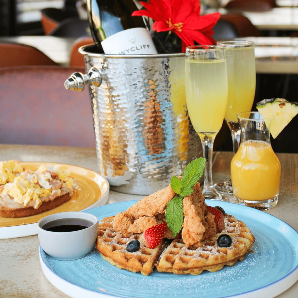 Chicken and waffles, scrambled eggs, mimosas, and champagne in a bucket on a table.