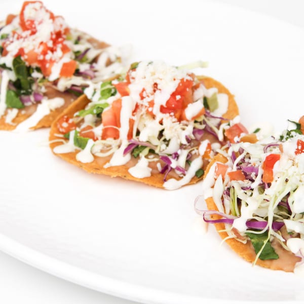 Three Mexican tostadas with shredded cabbage, tomatoes, and a creamy white sauce on a white plate.