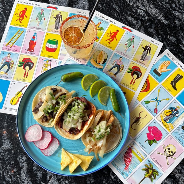 Mexican tacos and a margarita on a blue plate, surrounded by Loteria cards on a dark table.
