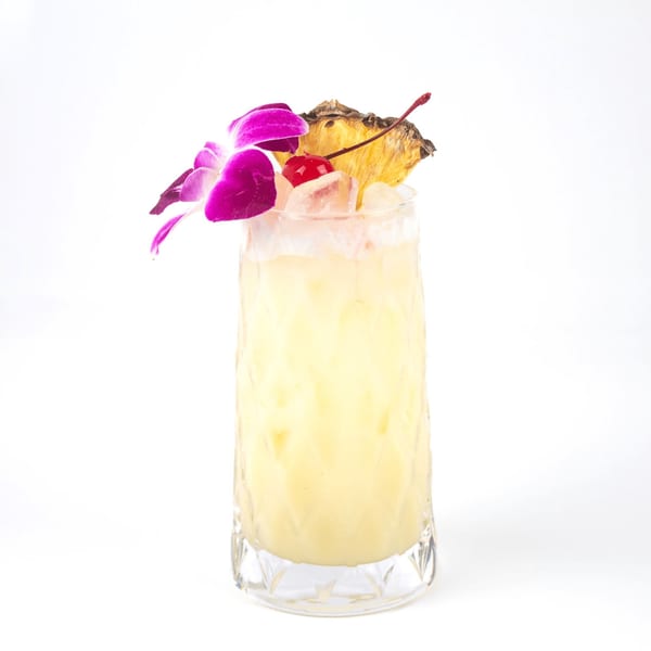 A tall glass of light yellow cocktail, garnished with a purple orchid, pineapple slice, and cherry.
