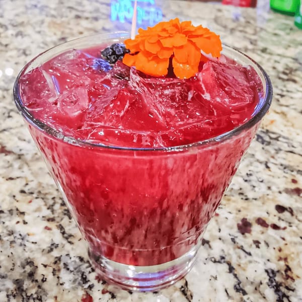 A vibrant red cocktail with ice, garnished with an orange flower and dark berries, on a speckled counter.