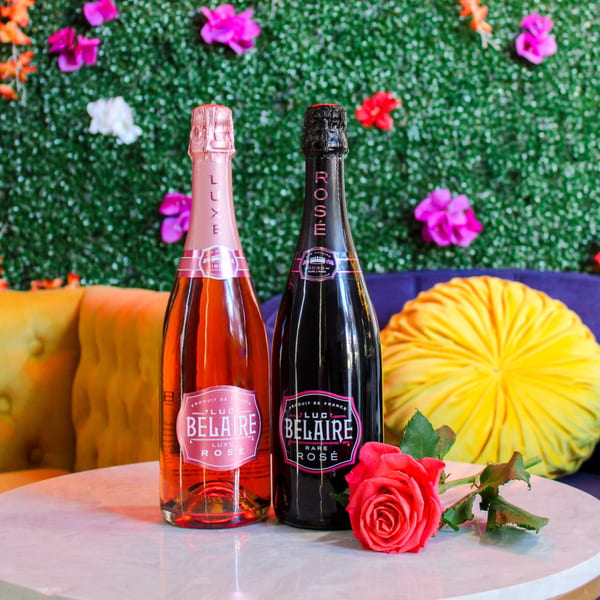Two bottles of Luc Belaire Rosé champagne and a red rose on a table with a floral wall.