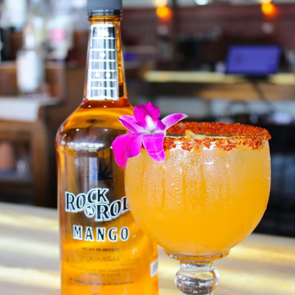 A Rock N Roll Mango tequila bottle next to a mango margarita with a chili-lime rim and orchid.