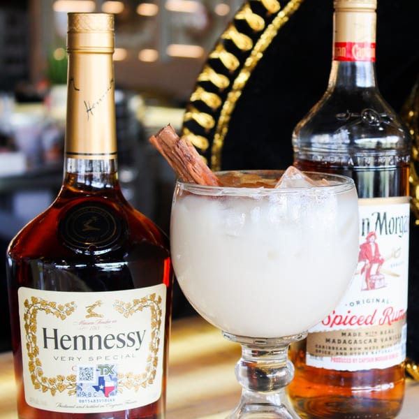 A creamy white cocktail with a cinnamon stick, flanked by bottles of Hennessy and Captain Morgan Spiced Rum.